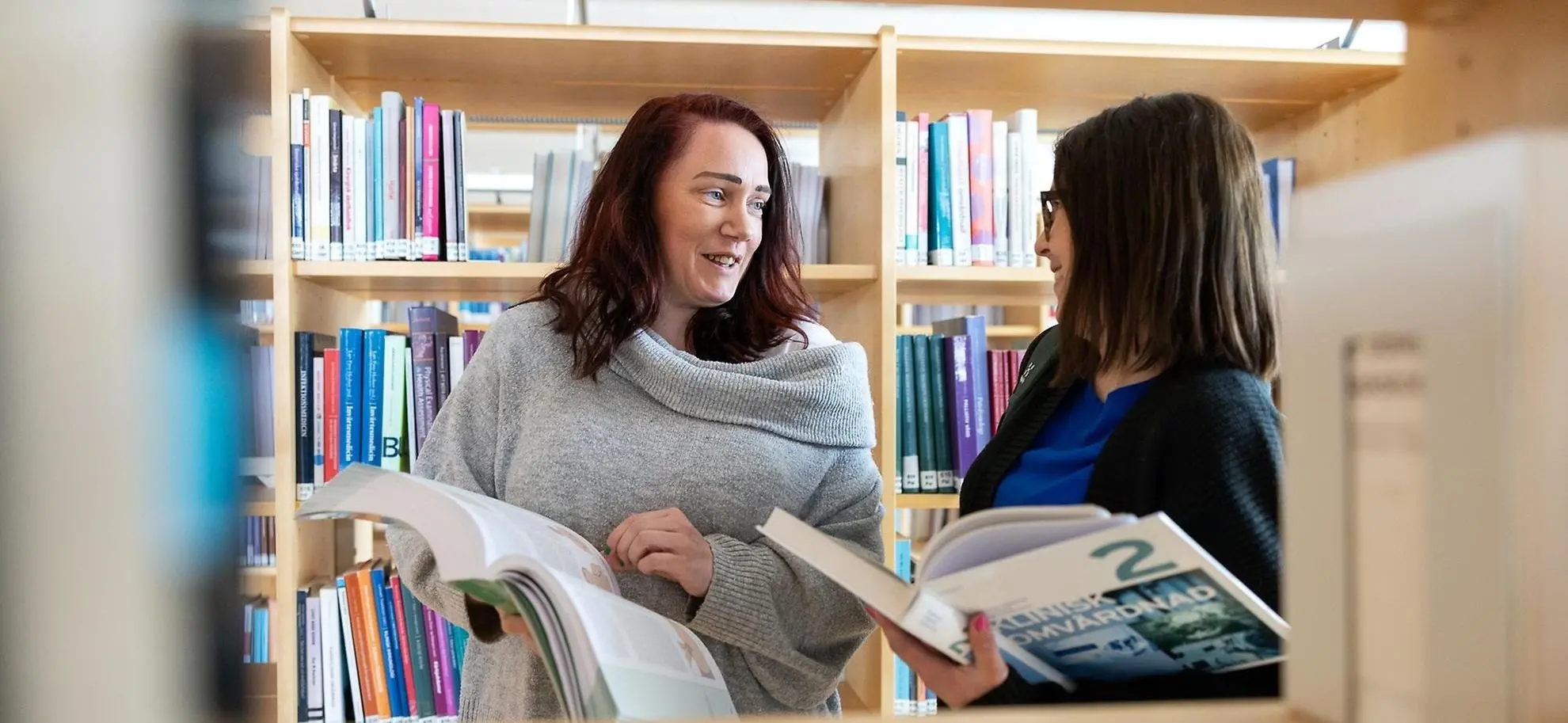 Två kvinnor står i ett bibliotek och håller i var sin bok