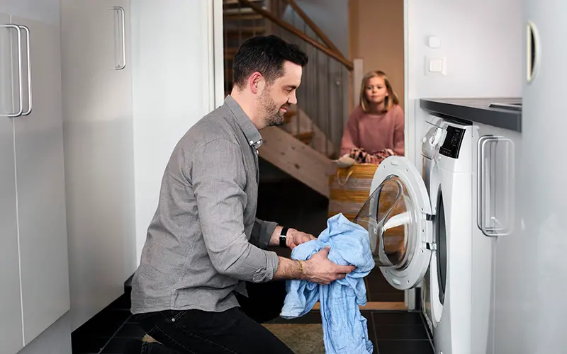En man lägger in tvätt i tvättmaskinen. En flicka i bakgrunden kommer med mer tvätt.