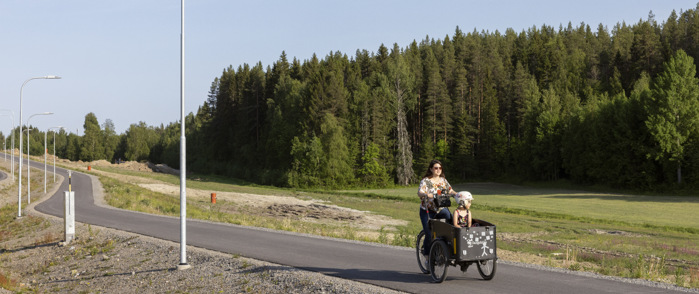 En vuxen cyklar på en elcykel längs en cykelväg. Det är sommar och i cykellådan frampå cykeln sitter ett barn.