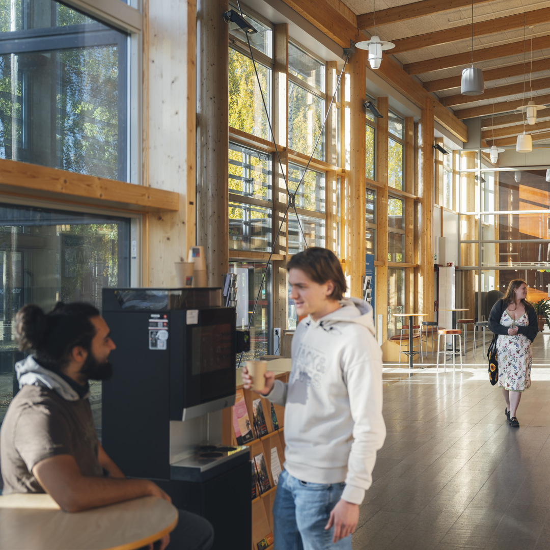 Några personer som står inne på campus. Två av personerna står vid kaffeautomaten och två går genom korridoren.