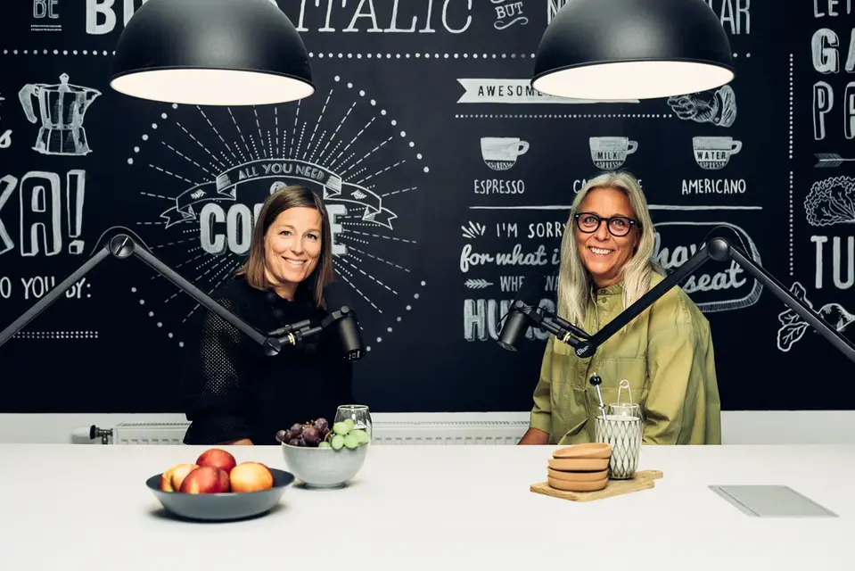 Maria Ekberg Brännström och Helena Renström sitter vid ett bord med radiomikrofoner och spelar in podden Skellefteå Stories
