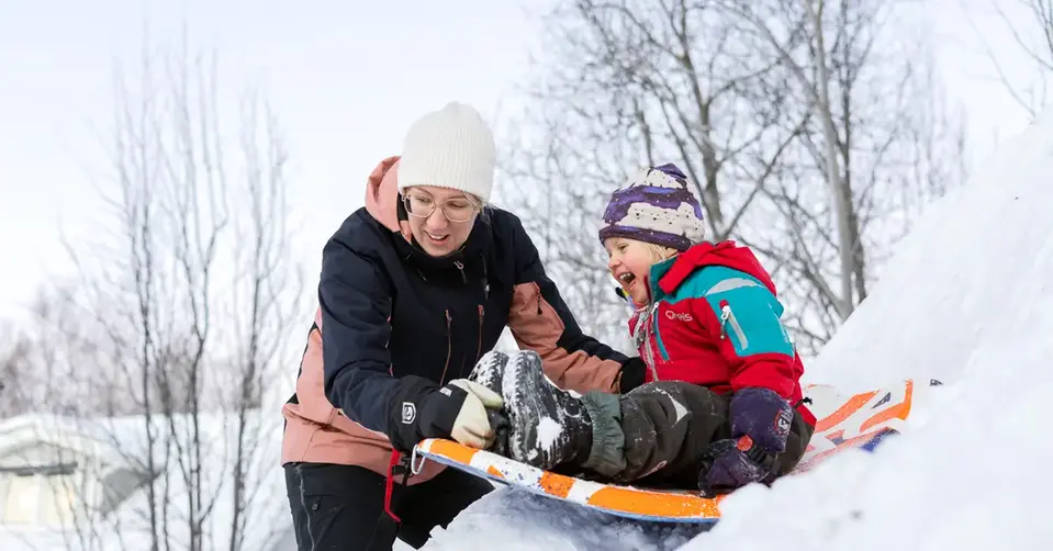 Sofia är ute och lekar i en snöhög med en av sina döttrar. Dottern sitter på en åkbräda, redo att glida nerför snöhögen.