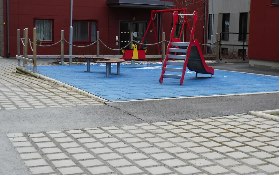 En lekplats med en blå och röd yta, en röd rutschkana och en bänk samt hårdgjord och jämn mark.